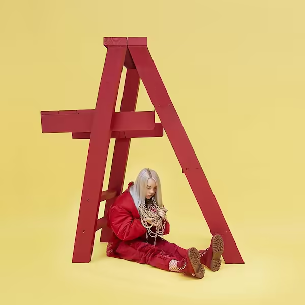 Eilish is seen sitting below a red ladder, holding jewelry that is around her neck.