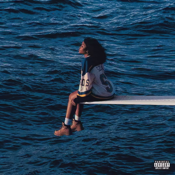 SZA sitting on the edge of a diving board in the middle of the water.