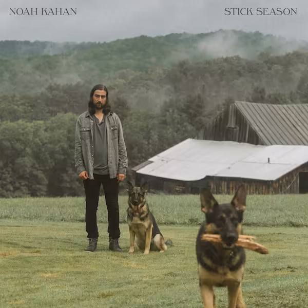 Kahan stands in the background of a barnyard setting. One German Shepherd sits beside Noah while the other Shepherd runs towards the foreground.
