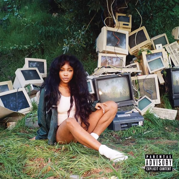 SZA sits on the grass in front of a pile of old monitors and keyboards.