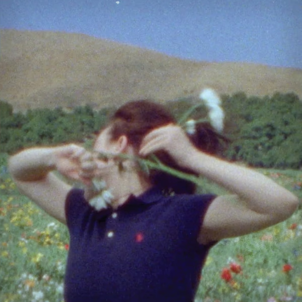 In a grainy image, Eilish in a blue short sleeve polo stands in a meadow of flowers, with a forest and mountain behind her. Her face is turned away from the camera and she is holding a white flower in her arms, raised towards the camera.